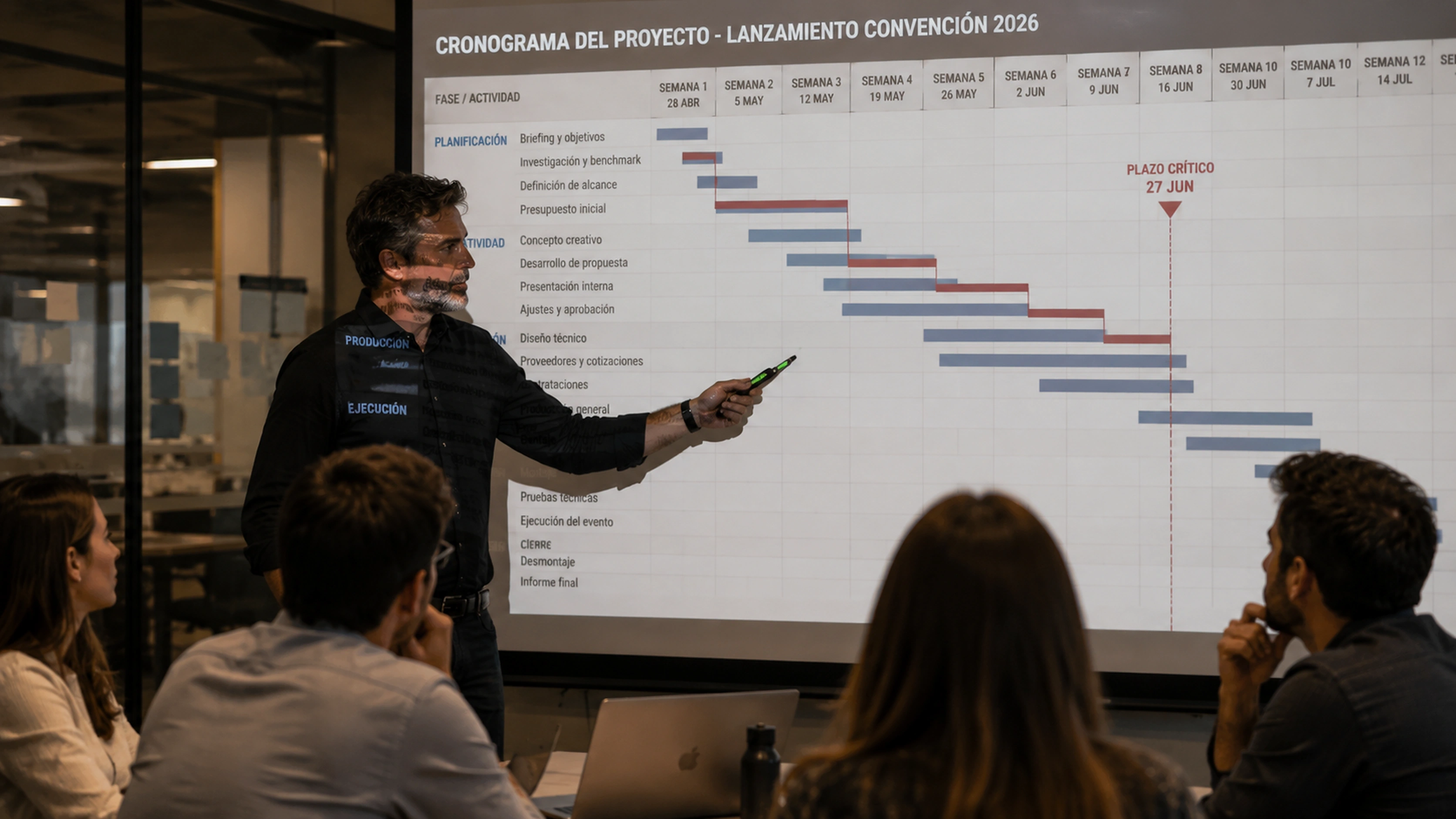 Productor senior presentando una carta Gantt al equipo y señalando un plazo crítico en la pantalla principal de una sala de reuniones.